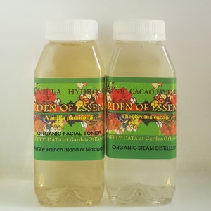 May include: Two clear glass bottles with green labels. The labels have white text that reads "Vanilla planifolia" and "Theobroma cacao". The bottles are labeled "Organic Facial Toner" and "Organic Steam Distilled".