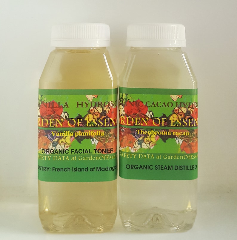 May include: Two clear glass bottles with green labels. The labels have white text that reads "Vanilla planifolia" and "Theobroma cacao". The bottles are labeled "Organic Facial Toner" and "Organic Steam Distilled".