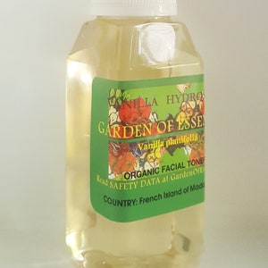 May include: A clear glass bottle with a white cap, containing a yellow liquid. The label is green with white text that reads "Vanilla Hydro Garden of Essen Vanilla planifolia Organic Facial Toner Read Safety Data at GardenOfEss Country: French Island of Made".