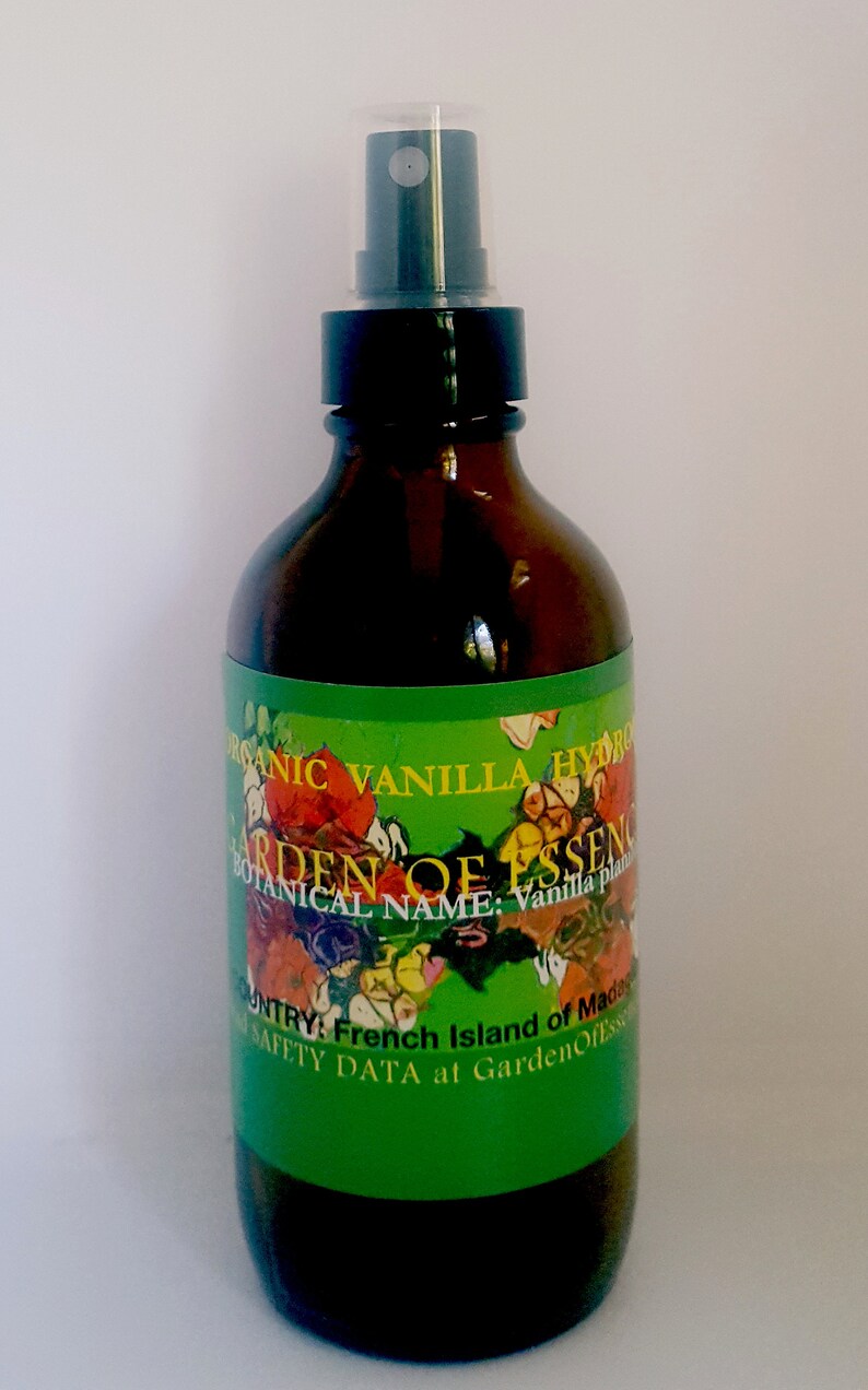 May include: A brown glass bottle with a black spray top and a green label. The label has a colorful floral design and the text "ORGANIC VANILLA HYDRO", "GARDEN OF ESSENCE", "BOTANICAL NAME: Vanilla plant", "COUNTRY: French Island of Madagascar", and "SAFETY DATA at GardenOfEssence".