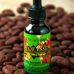 May include: A dark glass bottle with a black dropper top, labeled "Cacao Absol" and "Garden of Essence." The green label features colorful floral illustrations and text, surrounded by numerous brown cocoa beans. The bottle's origin is France.