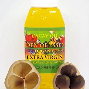 May include: A bottle of Cacay Oil with a white cap and a green and yellow label. The label reads "Cacay Oil, Garden of Essence, Organic, Extra Virgin". Two halves of a Cacay nut are in front of the bottle.