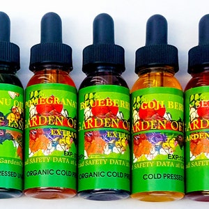 Puede incluir: Una fila de siete frascos cuentagotas de vidrio llenos de l&iacute;quidos de varios colores. Cada frasco tiene una tapa negra y una etiqueta verde con las palabras "Garden of Ess" y el nombre del aceite, como "Prickly Pear" y "Tamanu Oil".