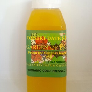 May include: A clear glass bottle with a white cap filled with a yellow liquid. The label reads "Desert Date Seed Oil, Garden of Ess, From the Sahara Desert, Extra Virgin, Organic Cold Pressed".