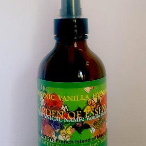 May include: A brown glass bottle with a black spray top. The label is green with a floral design and the text "ORGANIC VANILLA HYDRO", "GARDEN OF ESSENCE", "BOTANICAL NAME: Vanilla plant", "COUNTRY: French Island of Madagascar", and "SAFETY DATA at GardenOfEssence.com".