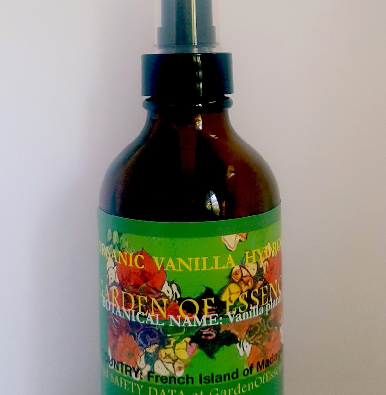 May include: A brown glass bottle with a black spray top. The label is green with a floral design and the text "ORGANIC VANILLA HYDRO", "GARDEN OF ESSENCE", "BOTANICAL NAME: Vanilla plant", "COUNTRY: French Island of Madagascar", and "SAFETY DATA at GardenOfEssence.com".