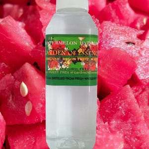 May include: A clear plastic bottle with a white cap, filled with a transparent liquid. The label reads "WATERMELON HYDROS GARDEN OF ESSENCE ORGANIC MELON FRUIT WATER ALCOHOL FREE". The background features bright red watermelon chunks.