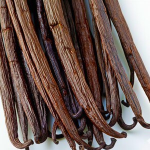 May include: A bunch of dried vanilla beans, brown in color, with a slightly wrinkled texture. The beans are arranged in a fan shape, with the tips of the beans pointing towards the top of the image.