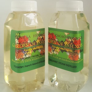 May include: Two clear glass bottles with white lids, filled with a light yellow liquid. The bottles are labeled "Garden of Essences" with the names of the essential oils, "Vanilla planifolia" and "Theobroma cacao", and the size, "8 oz". The labels also include the text "Organic Facial Tone", "Organic Steam Distilled", and "Read Safety Data at GardenOfEssences.com".