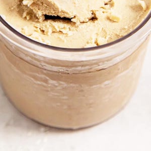 May include: A clear plastic container filled with light brown, textured food. The top portion of the food has a rough, uneven surface, while the lower portion appears smoother. The container is set against a white background.