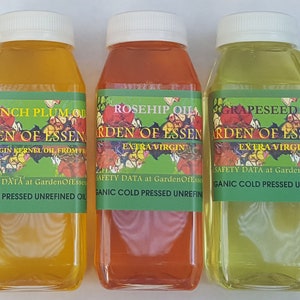 May include: Three bottles of oil with white caps and green labels. The labels read "French Plum Oil", "Rosehip Oil", and "Grapeseed Oil" with "Garden of Essen" and "Extra Virgin" text. The oils are amber, red-orange, and pale yellow.