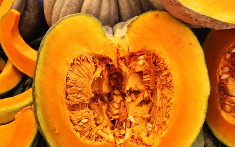 Puede incluir: Primer plano de una calabaza cortada por la mitad, que revela una pulpa de color naranja brillante y numerosas semillas. La superficie cortada muestra un interior texturizado. Se ven rodajas de calabaza en el fondo.