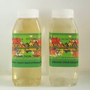 May include: Two clear glass bottles with green and white labels. The labels say "Garden of Essen" and list the ingredients as "Vanilla planifolia" and "Theobroma cacao". One bottle is labeled "Organic Facial Toner" and the other is labeled "Organic Steam Distilled".