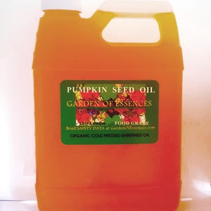 May include: An orange plastic jug with a white cap and handle, filled with pumpkin seed oil. A green label reads "PUMPKIN SEED OIL" and "GARDEN OF ESSENCES". The label also states "EXTRA VIRGIN FOOD GRABE" and "ORGANIC COLD PRESSED UNREFINED OIL".