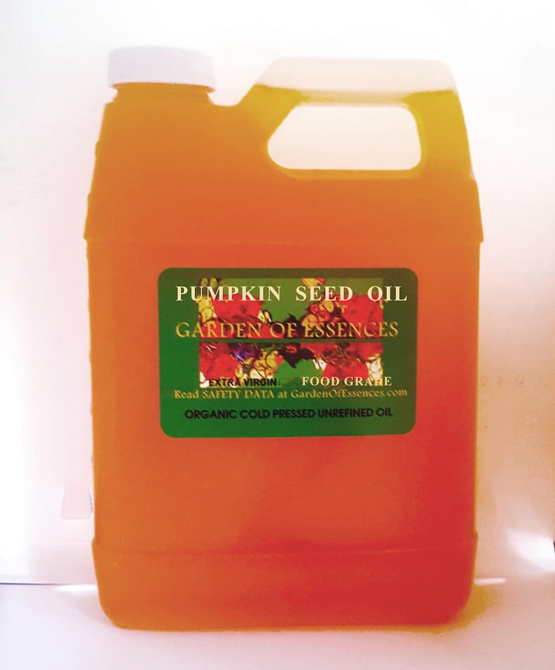 May include: An orange plastic jug with a white cap and handle, filled with pumpkin seed oil. A green label reads "PUMPKIN SEED OIL" and "GARDEN OF ESSENCES". The label also states "EXTRA VIRGIN FOOD GRABE" and "ORGANIC COLD PRESSED UNREFINED OIL".