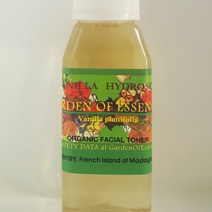May include: A clear glass bottle with a white cap filled with a light yellow liquid. The label reads "Vanilla planifolia Organic Facial Toner" and "Safety Data at GardenOfEssen" and "Country: French Island of Madagascar".