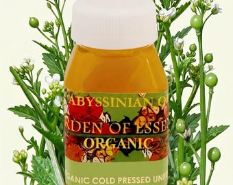 Könnte beinhalten: Eine kleine, transparente Flasche gefüllt mit goldener Flüssigkeit, mit einem weißen Deckel verschlossen. Das Etikett lautet "Abyssinian Oil Garden of Essence Organic Cold Pressed Unrefined". Umgeben von grünen Stielen, weißen Blüten und grünen Knospen.