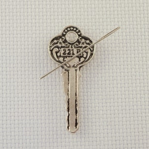 May include: A silver key with intricate details and the number "221" on the bow. A thin silver needle is placed diagonally across the key.