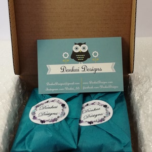 May include: Two teal blue gift boxes with white labels that say "Denkai Designs" are inside a brown cardboard box. The boxes are surrounded by white bubble wrap. A business card with an owl logo and the text "Denkai Designs" is visible in the background.