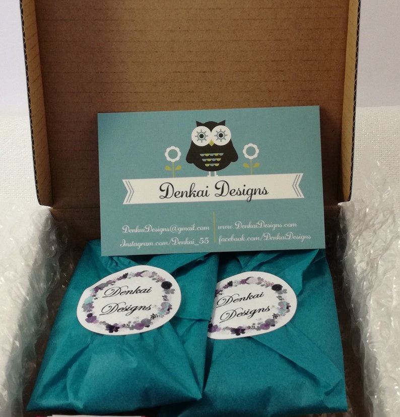 May include: Two teal blue gift boxes with white labels that say "Denkai Designs" are inside a brown cardboard box. The boxes are surrounded by white bubble wrap. A business card with an owl logo and the text "Denkai Designs" is visible in the background.