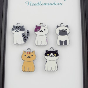 May include: Five silver cat needle minders with different colored details. The cats are sitting and have different colored eyes and fur. The text "Denkai Designs Needle minders" is at the top of the image and "www.DenkaiDesigns.com" is at the bottom.