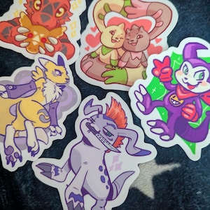 May include: A collection of five die-cut vinyl stickers featuring colorful cartoon creatures. The designs include a red dragon-like figure, two small creatures embracing with hearts, a yellow fox-like character, a purple imp, and a white creature with a blade.