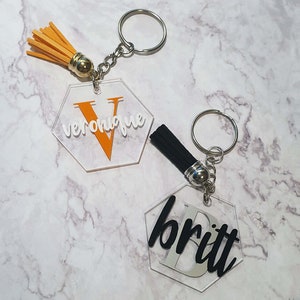 Op de afbeelding: Twee transparante, zeshoekige sleutelhangers van acryl met metalen ringen en kwastjes. Eén sleutelhanger heeft een zwart kwastje en de naam "Britt" in zwarte letters. De andere sleutelhanger heeft een oranje kwastje en de naam "Veronique" in witte letters.