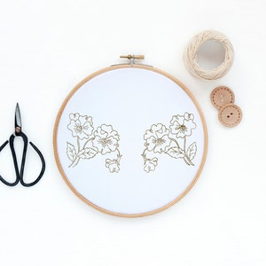 Peut inclure: Un cercle à broder en bois avec un tissu blanc tendu dessus. Le tissu est brodé d'un motif floral simple en fil brun. Le cercle est entouré d'une paire de ciseaux à broder noirs, d'une pelote de ficelle et de deux boutons bruns.