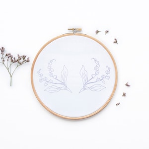 Op de afbeelding: Een witte borduurring met een eenvoudige lijntekening van twee lelietjes-van-dalen takken. De ring is op een witte achtergrond met verspreide gedroogde bloemen.