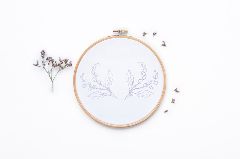Op de afbeelding: Een witte borduurring met een eenvoudige lijntekening van twee lelietjes-van-dalen takken. De ring is op een witte achtergrond met verspreide gedroogde bloemen.