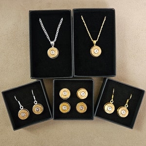 Könnte beinhalten: Drei Schmucksets mit Geschosshülsen. Das erste Set ist eine silberne Halskette mit einem Geschosshülsenanhänger. Das zweite Set ist eine goldene Halskette mit einem Geschosshülsenanhänger. Das dritte Set ist ein Paar Ohrringe mit Geschosshülsenanhängern.
