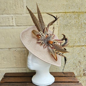 May include: A beige hat with a wide brim and a feather embellishment. The feathers are brown, tan, and white, and they are arranged in a cluster on the top of the hat.