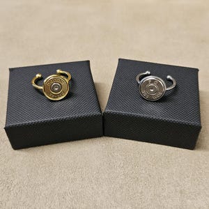 Puede incluir: Dos anillos, cada uno con una vaina de bala como pieza central. Uno tiene un acabado dorado con el texto "norma 22-250". El otro tiene un acabado plateado con el texto "F.C. 308 WIN". Ambos se muestran en cajas negras.