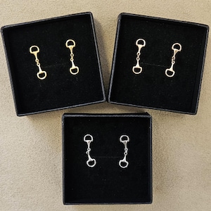 May include: Three pairs of earrings presented in black boxes. Two pairs are gold-coloured, and one pair is silver-coloured. Each earring features a small, detailed design resembling a horse bit, with a circular ring at each end connected by a chain.