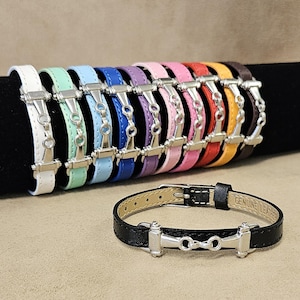 May include: A selection of leather bracelets in a spectrum of colours, such as white, green, blue, purple, pink, red, yellow, and brown. Each bracelet has a silver-toned metal bit design. A black leather bracelet with a silver bit is also shown.