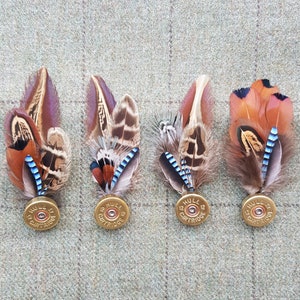 May include: Four pheasant feather and shotgun shell brooches. Each brooch features a cluster of brown, tan, and blue feathers with a gold shotgun shell base. The shotgun shells are labeled "12 Gauge Cartridge".