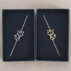 May include: Two small boxes with black velvet interiors, each containing a silver and gold origami fox bracelet. The bracelets are delicate and feature a geometric fox design.