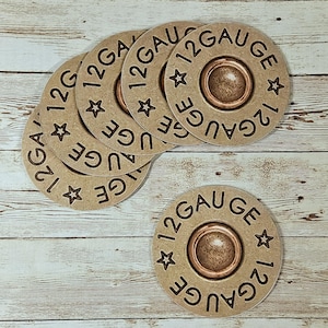 May include: Six circular coasters with a tan, metallic finish. Each coaster features the text "12 GAUGE" around the edge, with star accents. The centre of each coaster has a metallic brown circle. The coasters are made of cardboard.