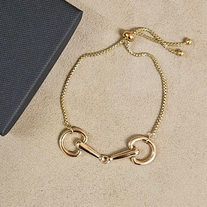 May include: A gold-coloured bracelet featuring a central horse bit motif connected to a delicate box chain. The bracelet includes an adjustable sliding mechanism with two small gold beads at the ends, allowing for a customisable fit on the wrist.