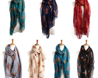 Puede incluir: Colección de bufandas ligeras en varios colores, cada una adornada con un patrón de zorro repetido. Las bufandas se exhiben en maniquíes, mostrando las diferentes opciones de color: verde azulado, azul marino, rojo, gris, beige, turquesa y negro.
