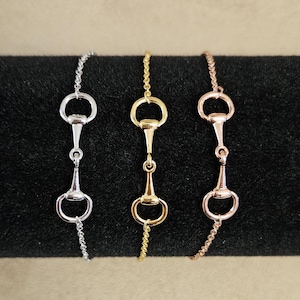 May include: Three equestrian-themed bracelets in silver, gold, and rose gold. Each bracelet features a horse bit design as the central element, connected to a delicate chain. The bracelets are displayed against a dark background.