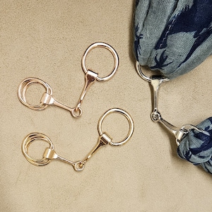 May include: Three gold-toned metal horse bit charms with a ring on one end and a hook on the other. The charms are on a light brown surface.