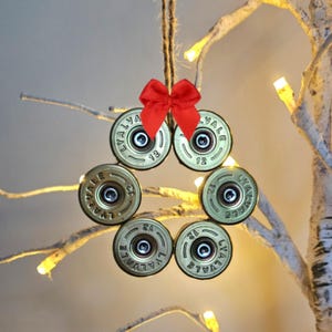 May include: A festive Christmas wreath ornament made from six brass shotgun shell casings, arranged in a circular shape. A small red bow adorns the top. The shells are suspended by twine and are suitable for a Christmas tree.