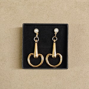 Puede incluir: Un par de pendientes de oro con un acento de perla. Los pendientes tienen forma de herradura con un pequeño bucle en la parte superior.