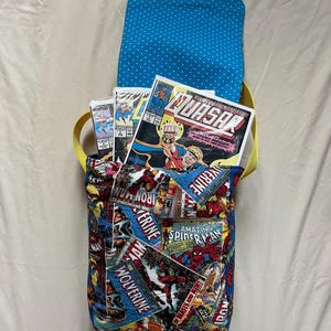 May include: A handmade comic book-themed messenger bag. The bag features a vibrant collage of comic book covers, including "Iron Man," "Wolverine," and "Amazing Spider-Man." The interior lining is blue with white polka dots, and the strap is yellow.
