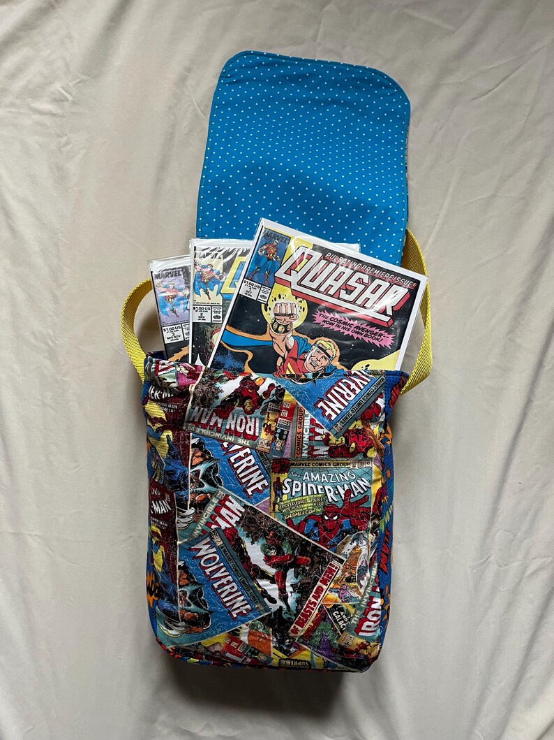 May include: A handmade comic book-themed messenger bag. The bag features a vibrant collage of comic book covers, including "Iron Man," "Wolverine," and "Amazing Spider-Man." The interior lining is blue with white polka dots, and the strap is yellow.