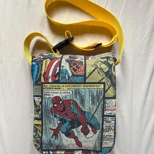May include: A rectangular comic book-themed crossbody bag with a yellow strap. The bag features panels with Spider-Man and other Marvel characters. The text "AND, SPEAKING OF OUR FRIENDLY NEIGHBORHOOD SPIDER-MAN... I DON'T MIND A LITTLE RAIN..." is visible.