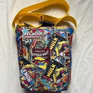 May include: A handmade, fabric crossbody bag featuring a collage of comic book covers, including Iron Man, Spider-Man, and Hulk. The bag has a yellow strap and a black buckle. The bag is approximately 8 inches tall.