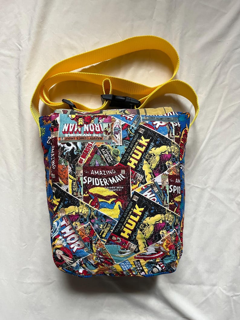 May include: A handmade, fabric crossbody bag featuring a collage of comic book covers, including Iron Man, Spider-Man, and Hulk. The bag has a yellow strap and a black buckle. The bag is approximately 8 inches tall.