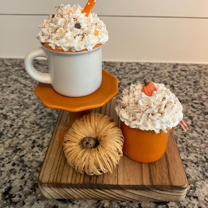 May include: A white mug and an orange mug topped with white whipped cream, cinnamon sprinkles, and small orange accents. The white mug sits on an orange pedestal, accompanied by a woven raffia pumpkin on a wooden base.